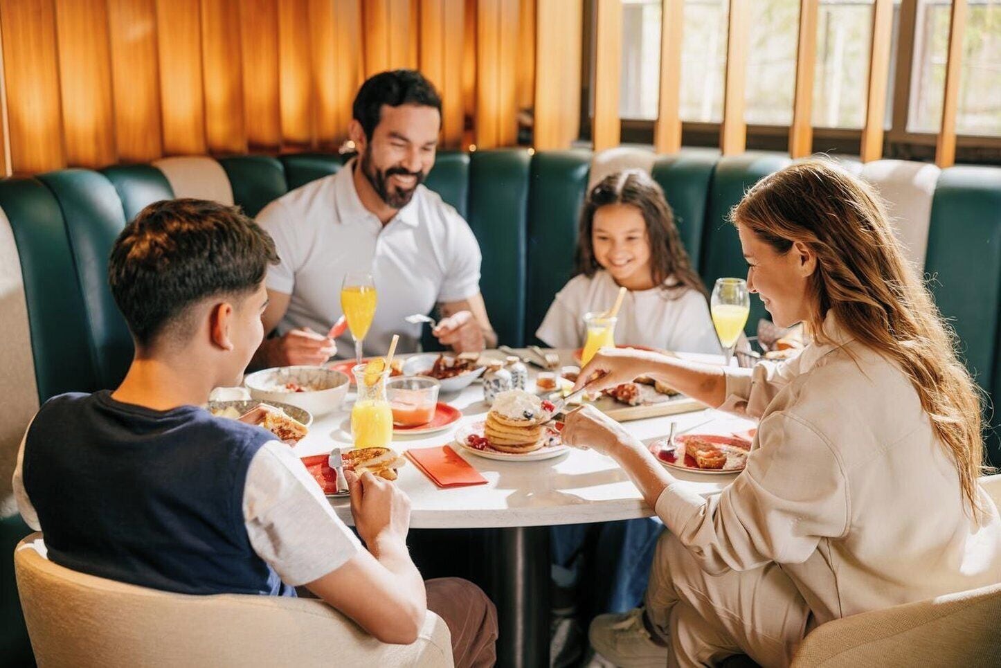 Una familia cenando en un restaurante con asientos tipo cabina, una mesa con comida para el desayuno, y una ventana con luz natural.