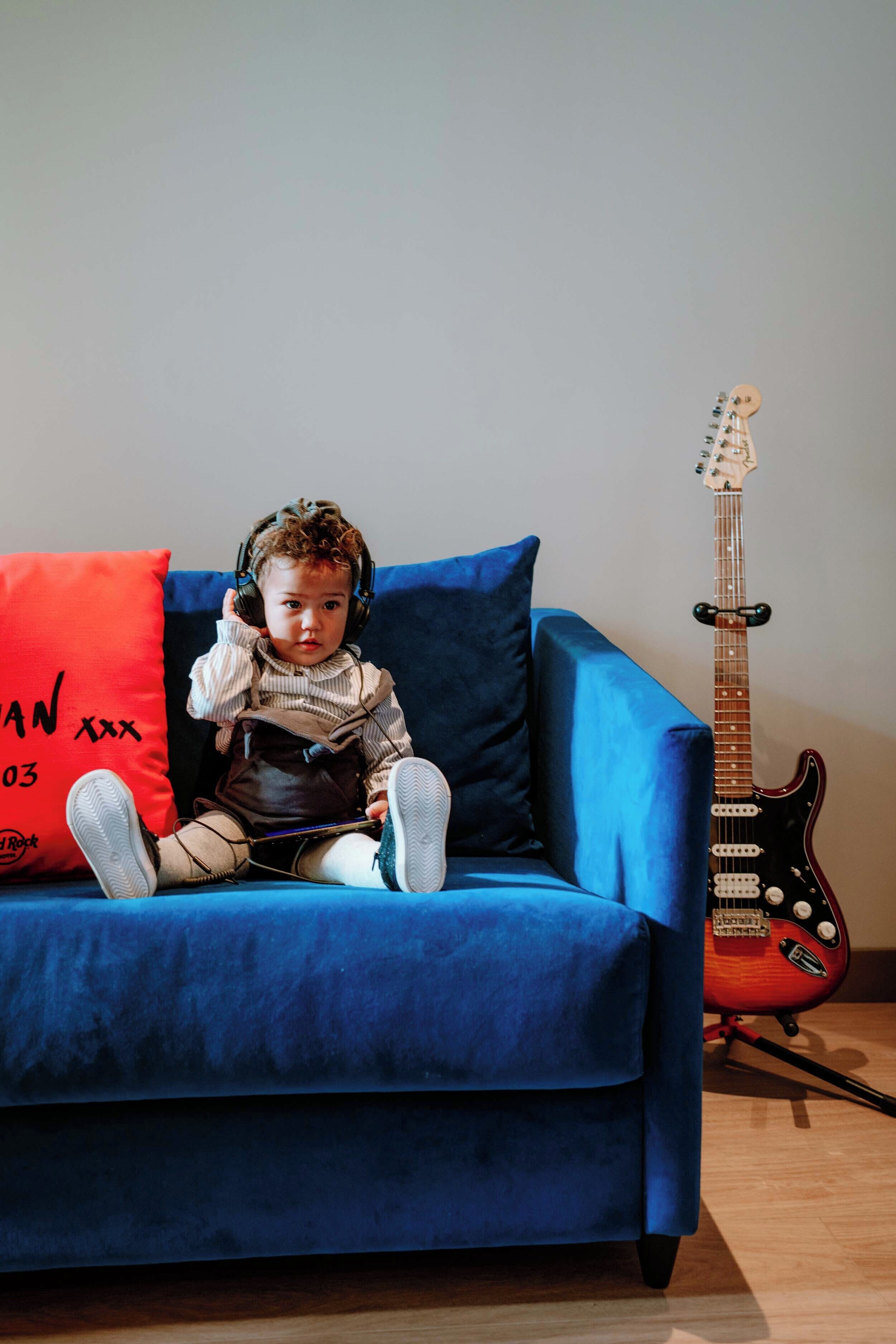 A child sitting on a blue sofa with headphones, a red pillow, and a guitar in the background.