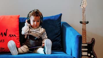 Un enfant est assis sur un canapé bleu, portant un casque audio, avec un coussin rouge et une guitare en arrière-plan.