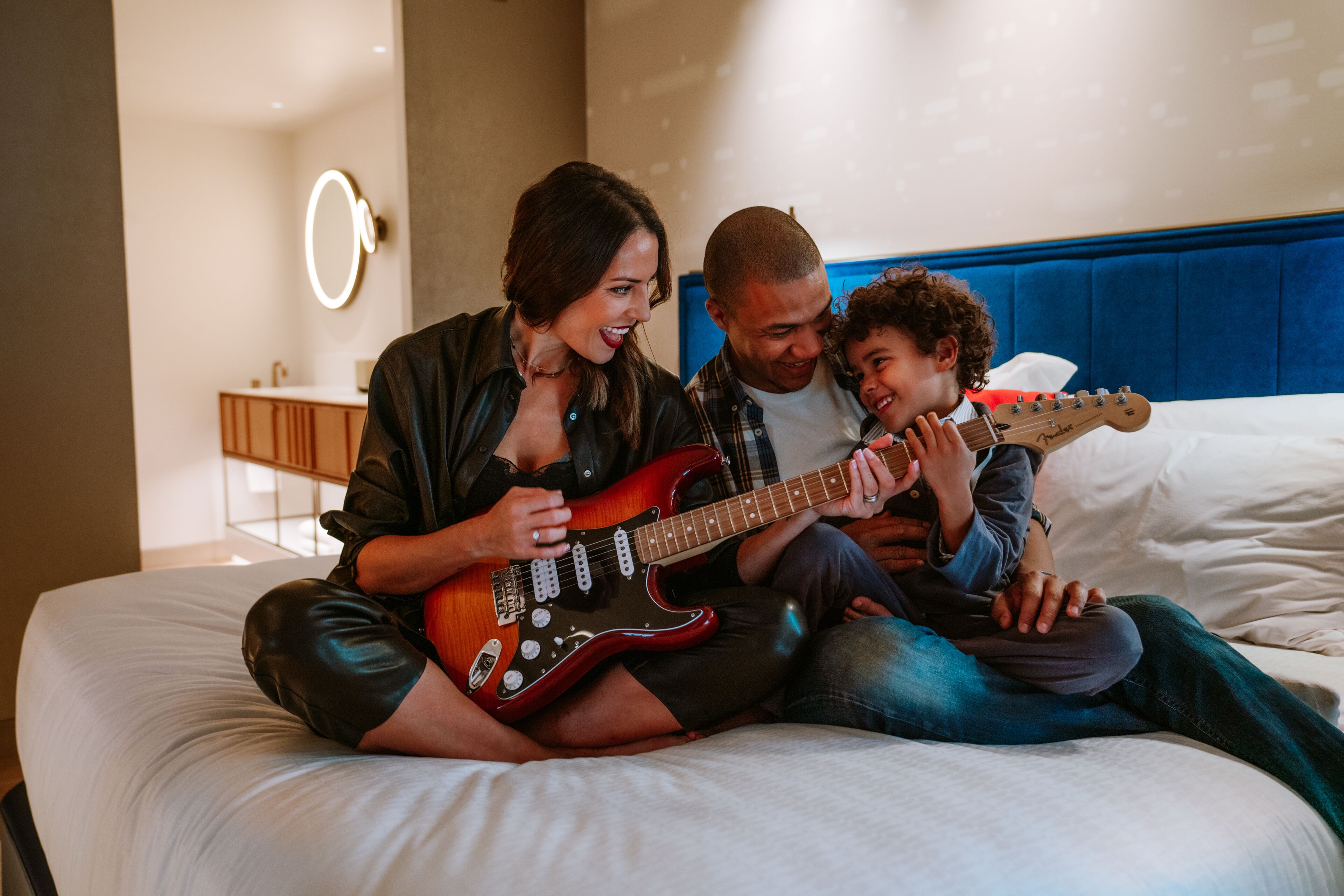 Una familia sentada en una cama en una habitación de hotel con un cabecero azul, un espejo y una mesita de noche con una lámpara.