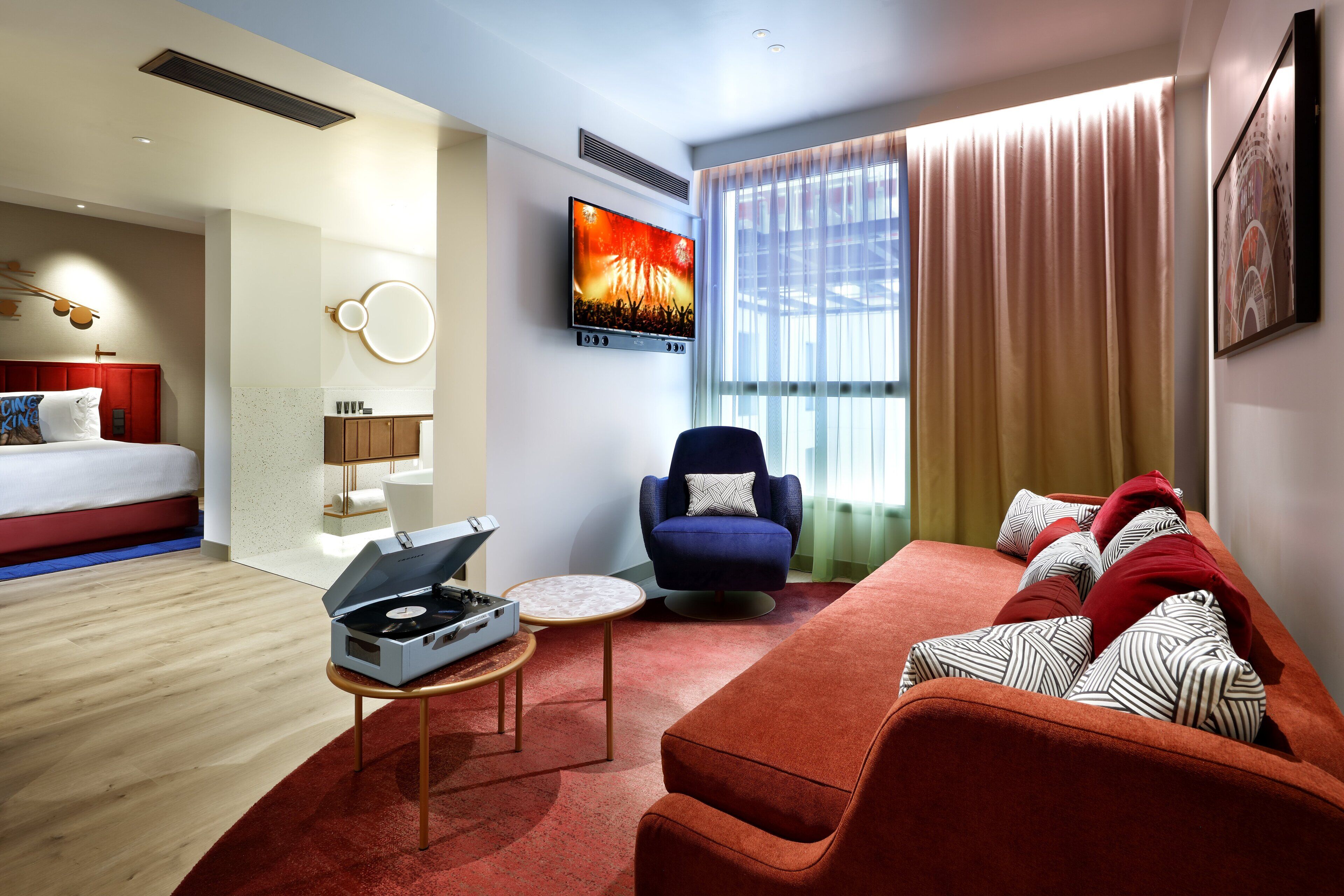 Una habitación de hotel moderna con una cama, un sofá rojo, un sillón azul, un televisor de pantalla plana y un reproductor de DJ.