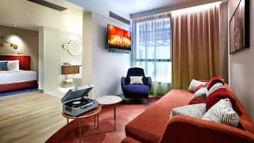Una habitación de hotel moderna con una cama, un sofá rojo, un sillón azul, un televisor de pantalla plana y un reproductor de DJ.