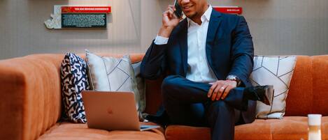 A man in a suit sitting on an orange couch with a laptop, talking on a phone. There are framed pictures and a mannequin on the wall behind him.