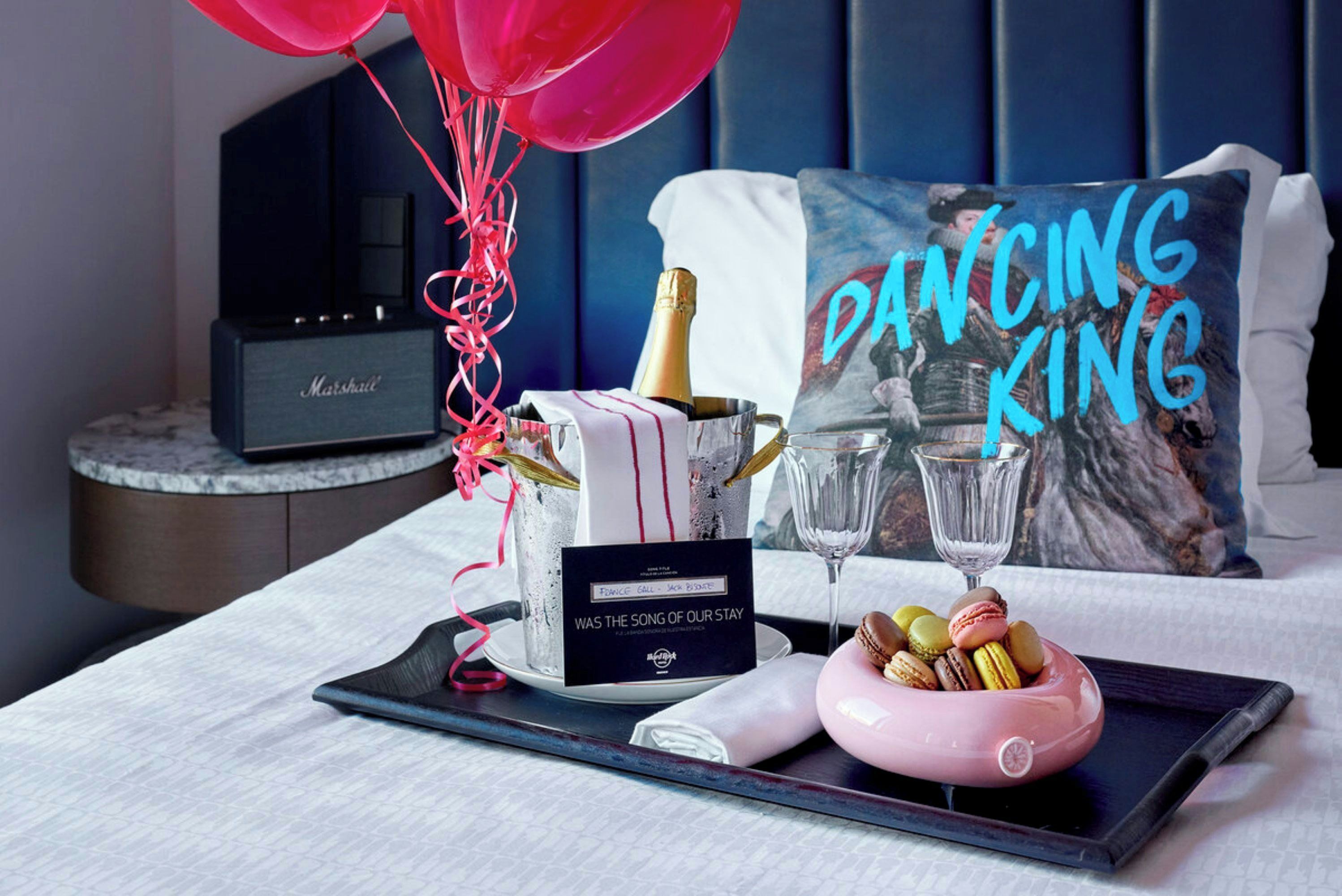 Ein Hotelzimmer mit einem Bett, einem Nachttisch mit einem Marshall-Lautsprecher, einer Champagnerflasche, Gläsern, einer Karte und einer Schale mit Macarons.
