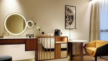 Habitación de hotel moderna con un espejo redondo, una cafetera, una mesita pequeña, una silla y una obra de arte enmarcada en la pared.