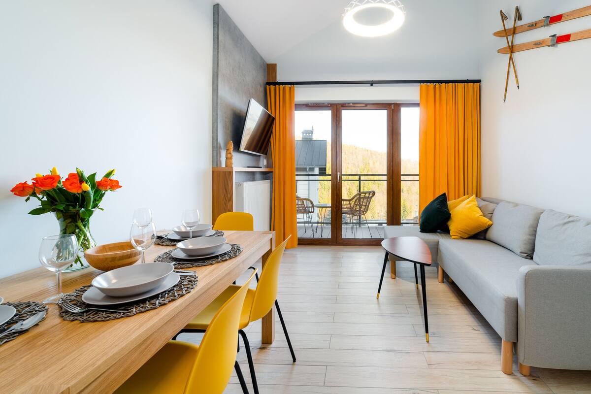 Un salon moderne avec un canapé gris, des fauteuils jaunes, une table à manger en bois avec des assiettes et des verres, et un vase rempli de fleurs.