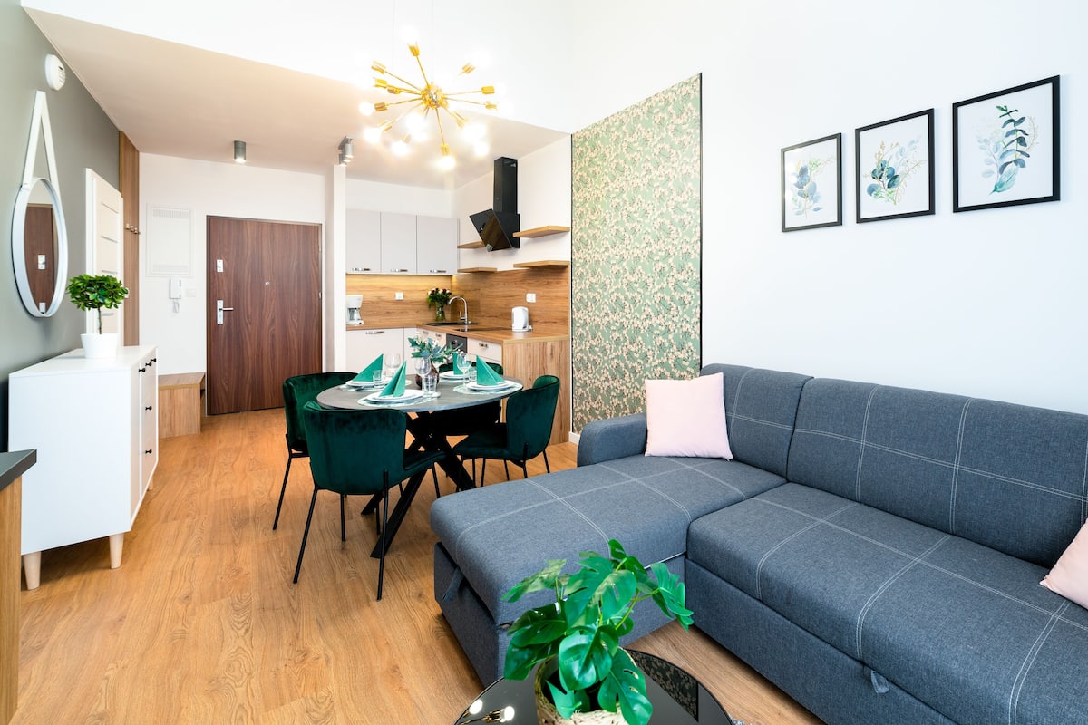 Un salon moderne avec un canapé gris, une table à manger dressée pour quatre personnes et un coin cuisine à l’arrière-plan.
