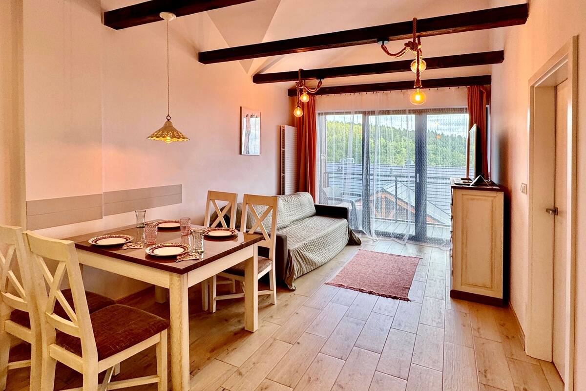 Un coin repas accueillant, avec une table en bois dressée pour quatre personnes, un canapé et une vue sur l’extérieur grâce à une grande fenêtre.