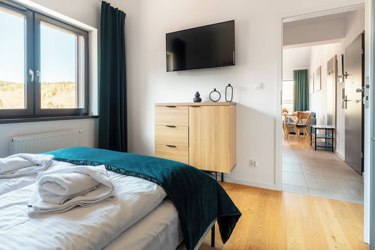 Une chambre à coucher avec un lit, un téléviseur fixé au mur, une commode en bois et une vue sur une forêt par la fenêtre.