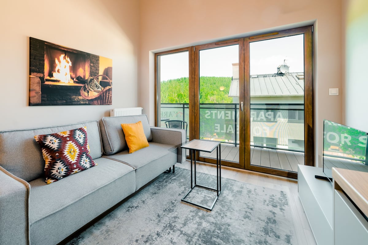 Un salon moderne avec un canapé gris, des coussins colorés, une petite table et une grande porte-fenêtre coulissante donnant sur un balcon avec vue sur la verdure et les bâtiments.