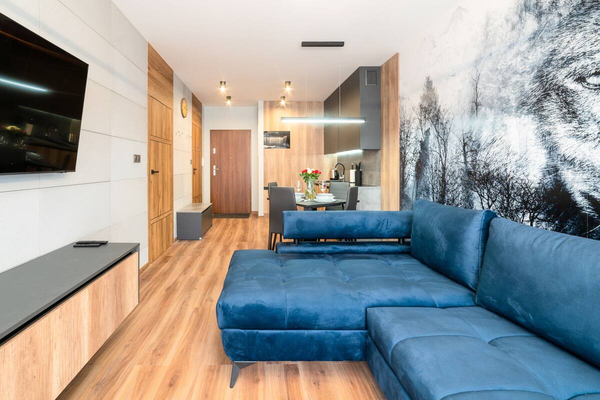 Un salon moderne avec un canapé bleu, un plancher en bois, un téléviseur à écran plat fixé au mur et un coin repas à l’arrière-plan.