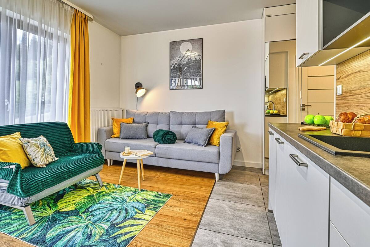 Un salon moderne avec un canapé vert, un canapé gris et une petite table sur laquelle se trouve un livre. On y trouve également un coin cuisine avec un plan de travail en bois et un évier.