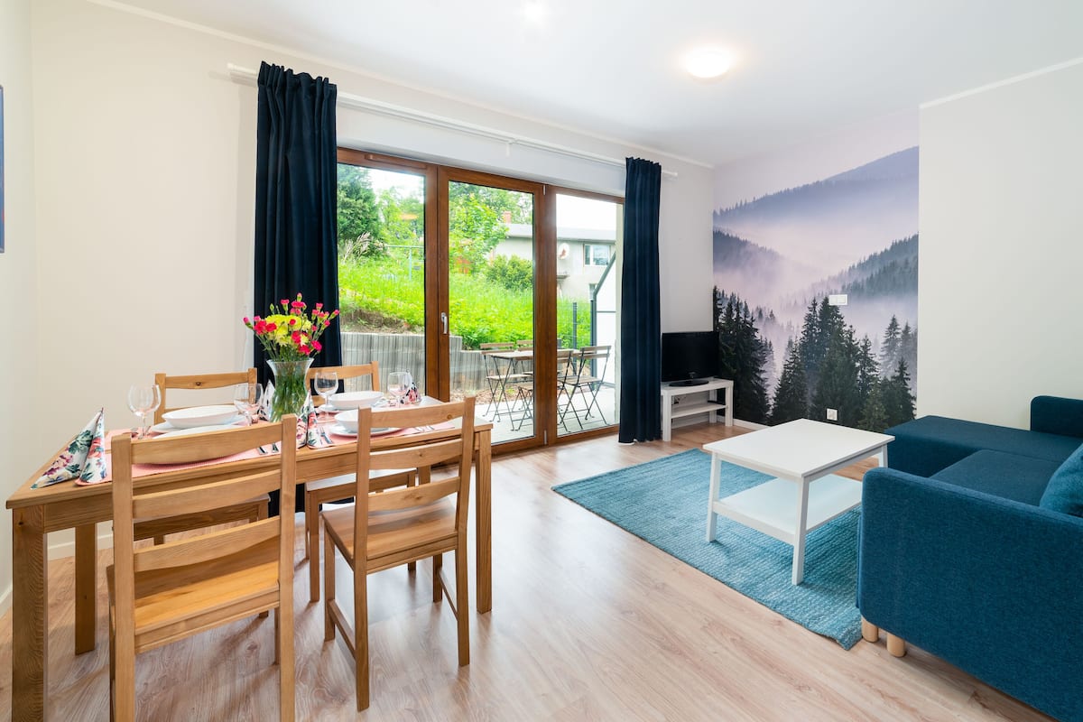 Un salon comprenant un coin repas, un canapé, un téléviseur et une fresque représentant une forêt.