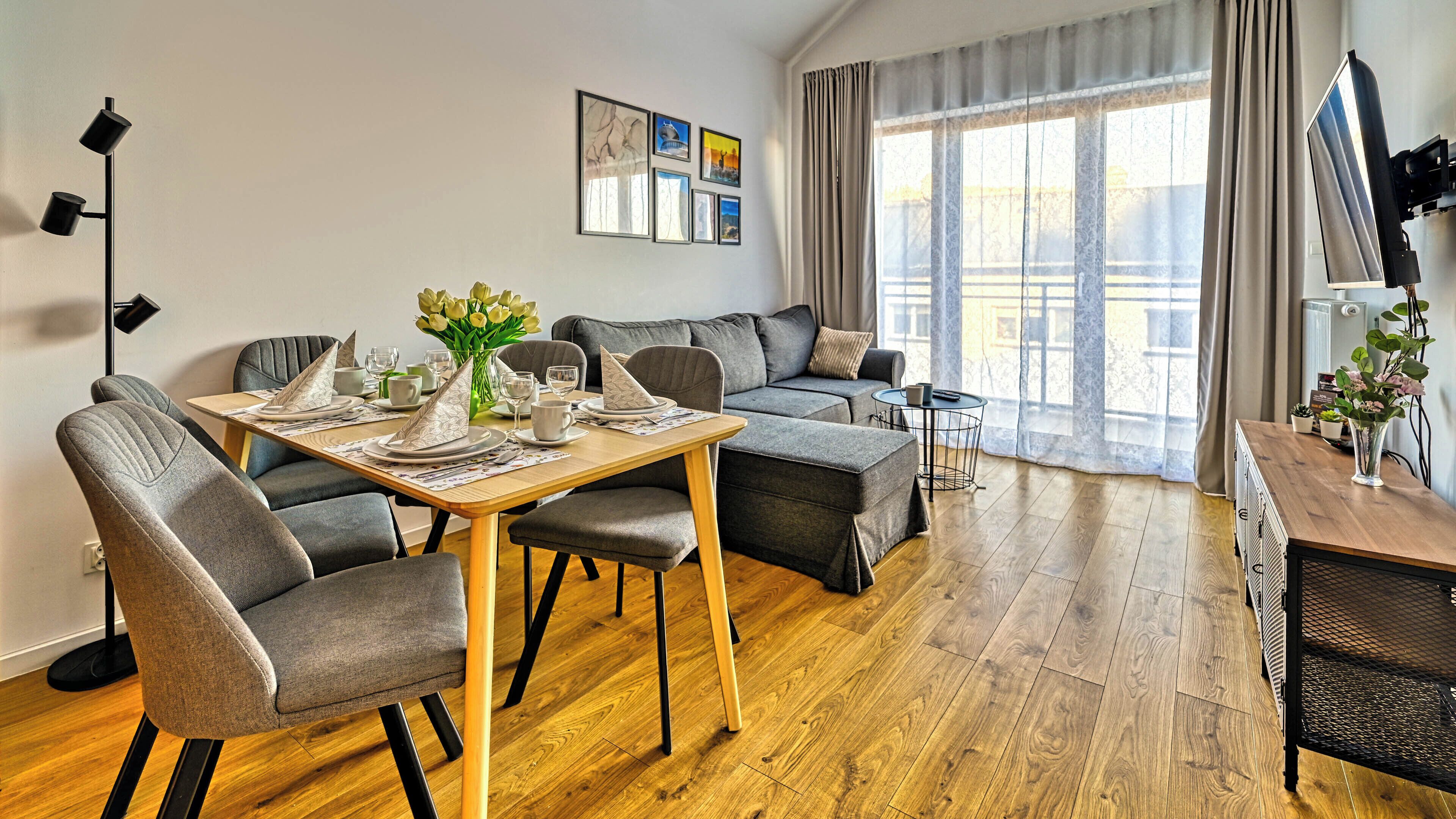A modern living room with a dining area, grey sofas, a wooden dining table, and a large window with curtains.