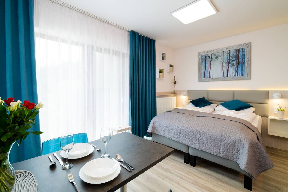 Une chambre d’hôtel moderne, avec un lit soigneusement fait, une table à manger dressée pour deux personnes et un vase rempli de fleurs.