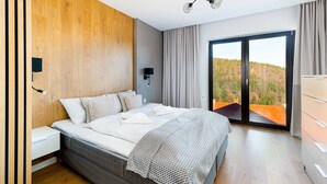 A modern bedroom with a large bed, a nightstand, a wall-mounted light, and a view of a forest through a sliding glass door.