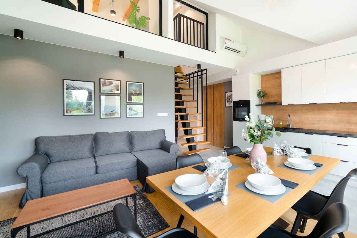 Un appartement moderne comprenant un coin repas, un canapé gris, une table à manger en bois pour quatre personnes et une cuisine avec des meubles blancs et un plan de travail noir.