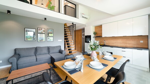 Un apartamento moderno con comedor, un sofá gris, una mesa de comedor de madera para cuatro comensales, y una cocina con alacenas blancas y encimera negra.