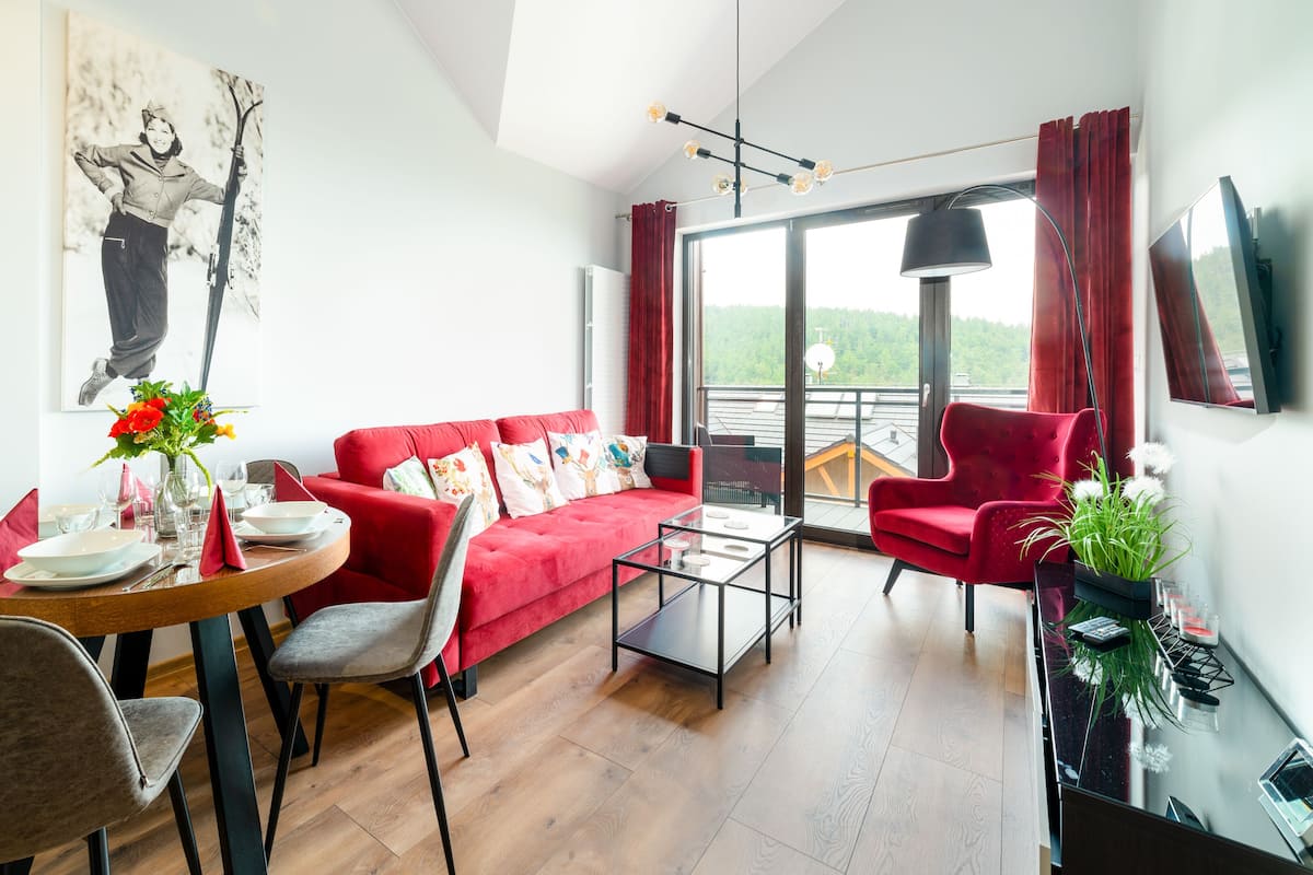Un salon moderne avec un canapé rouge, une table à manger avec des chaises, une table basse en verre et un téléviseur fixé au mur.
