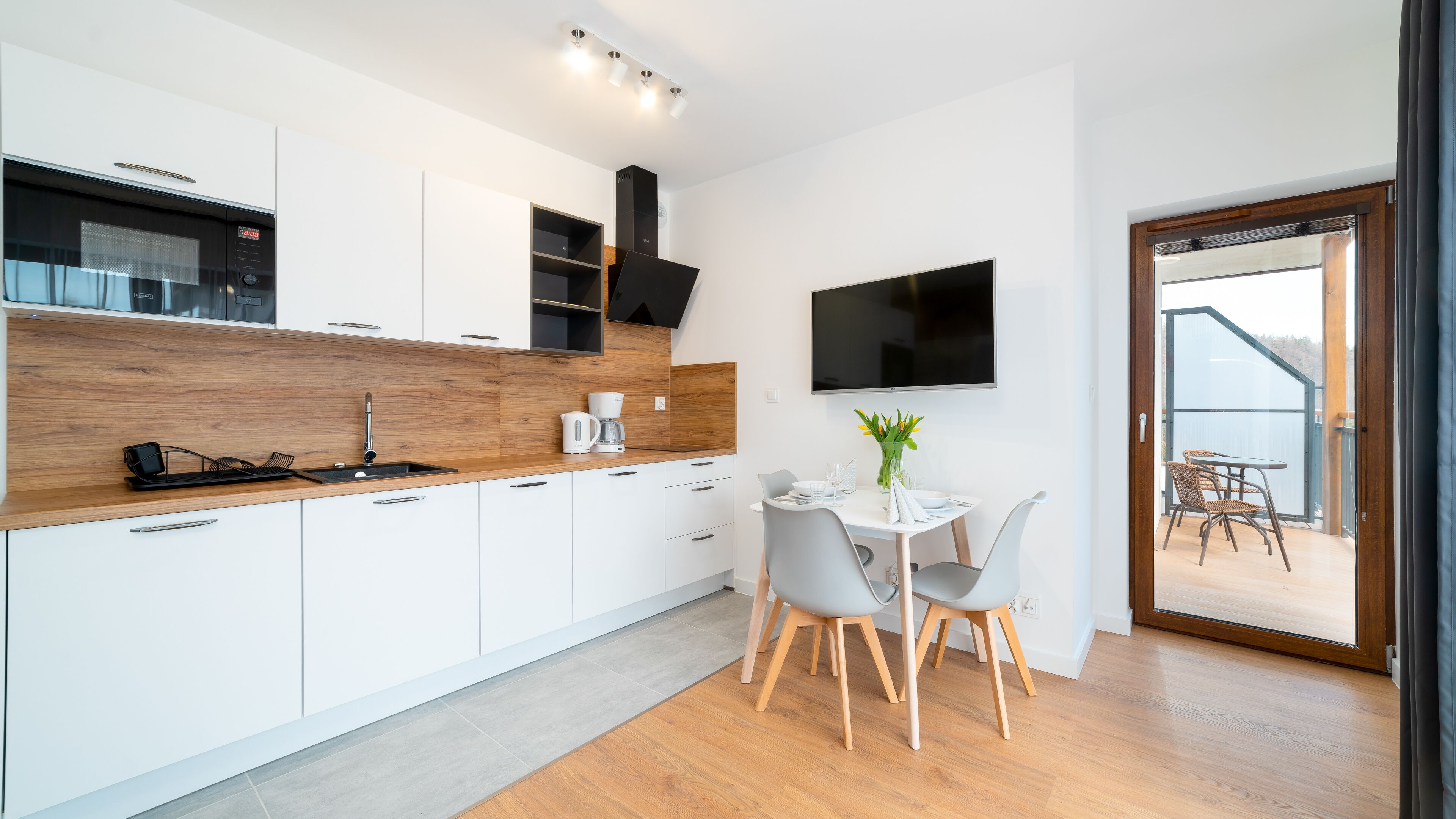 A modern kitchen with white cabinets, a wooden countertop, and a built-in oven. There is a dining area with a table and chairs, a TV mounted on the wall, and a view of the outdoors through a glass door.