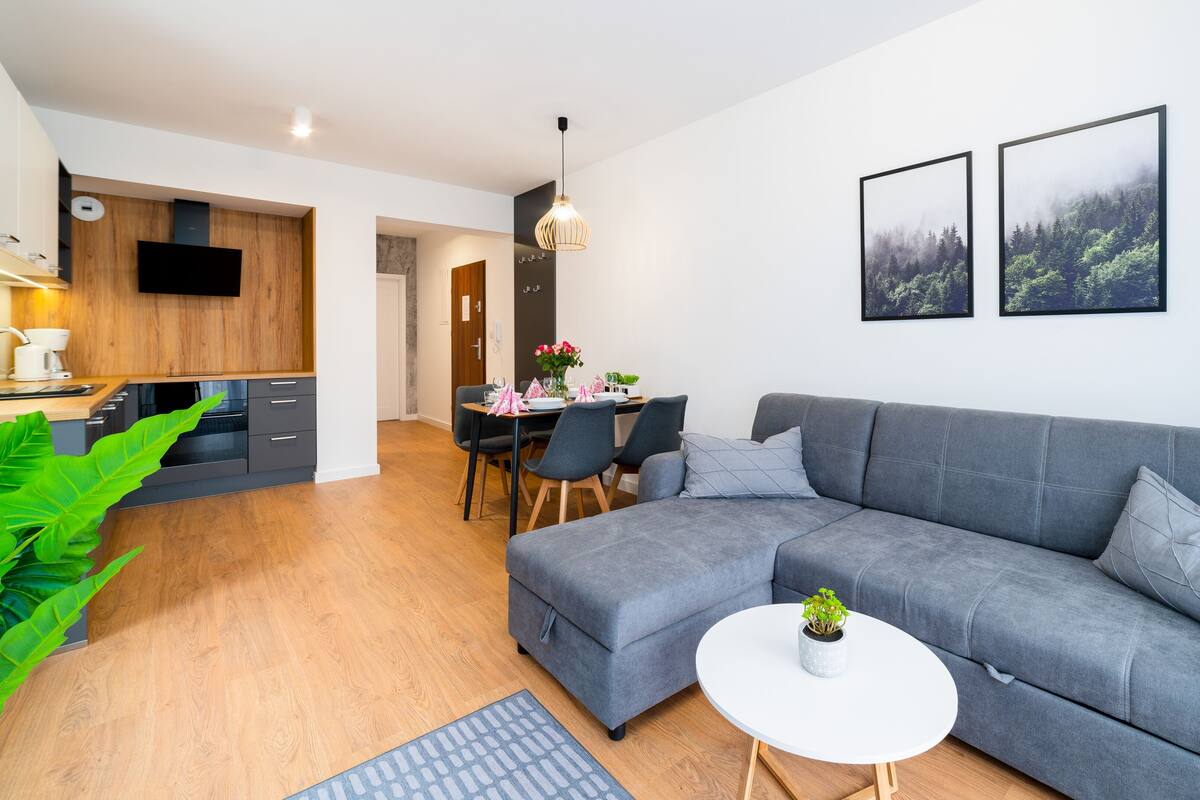 Un salon moderne avec un canapé gris, un coin repas comprenant une table et des chaises, et une cuisine dotée de meubles en bois et d’un téléviseur à écran plat.