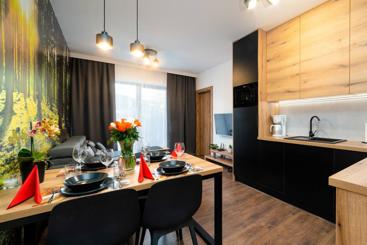 Une cuisine moderne avec des meubles en bois, un réfrigérateur noir et un coin repas avec une table dressée pour deux personnes.
