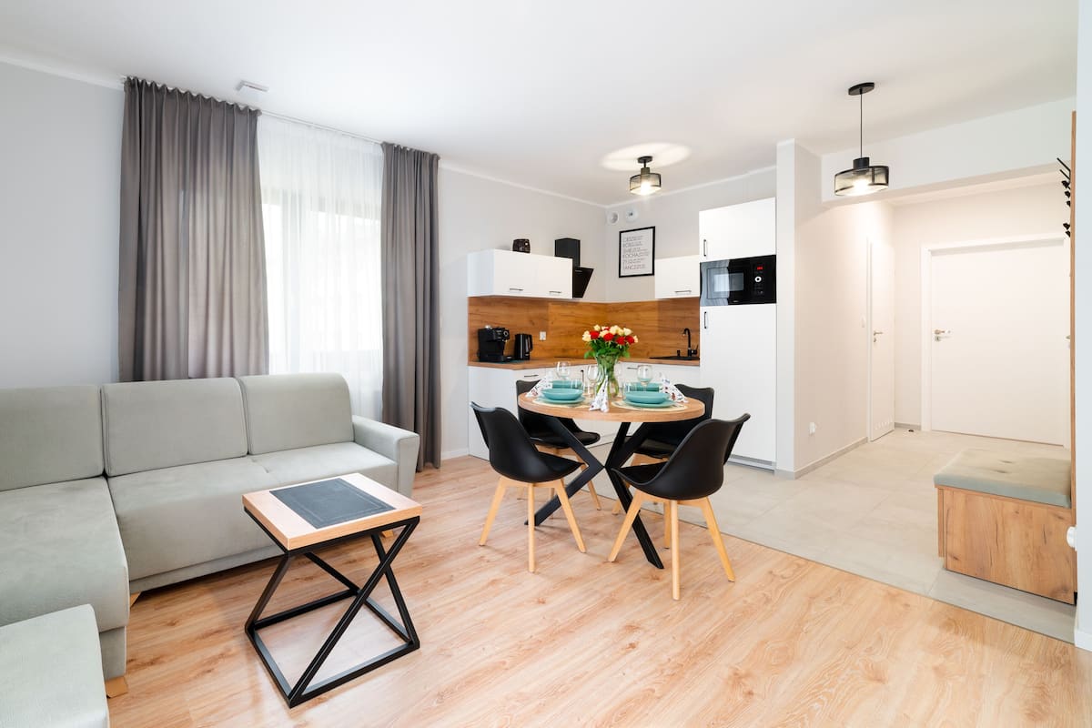 Un appartement moderne avec un canapé gris, une table à manger avec des chaises, une petite table d’appoint en bois et un coin cuisine équipé d’un four à micro-ondes.