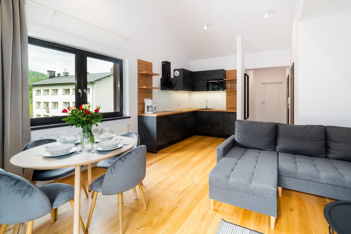 Un salon moderne avec un canapé gris, une table à manger avec des chaises et un coin cuisine doté de meubles sombres et d’un plan de travail en bois.