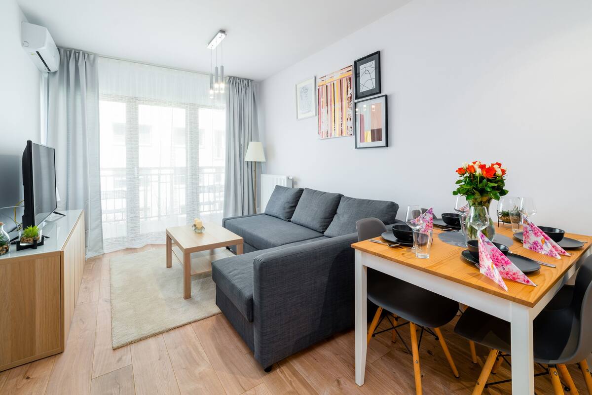 Un salon moderne avec un canapé gris, une table à manger en bois dressée pour deux personnes et un téléviseur posé sur un meuble en bois.