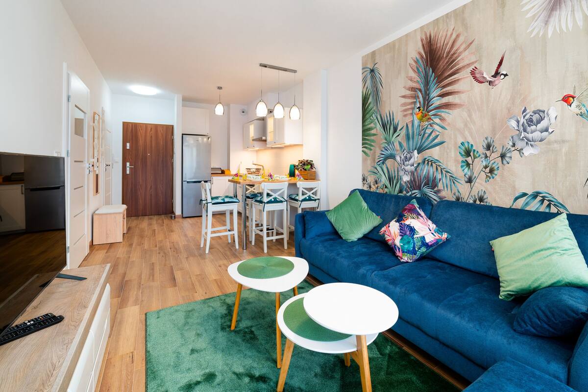 Un salon moderne avec un canapé bleu, des coussins verts et une fresque murale aux motifs tropicaux.