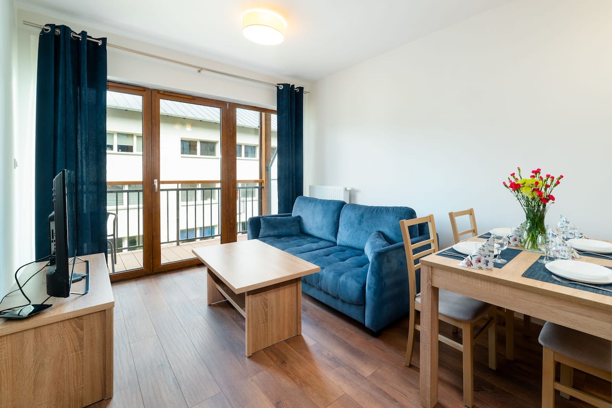 Un salon moderne avec un canapé bleu, une table à manger en bois dressée pour deux personnes et un balcon agrémenté de rideaux.