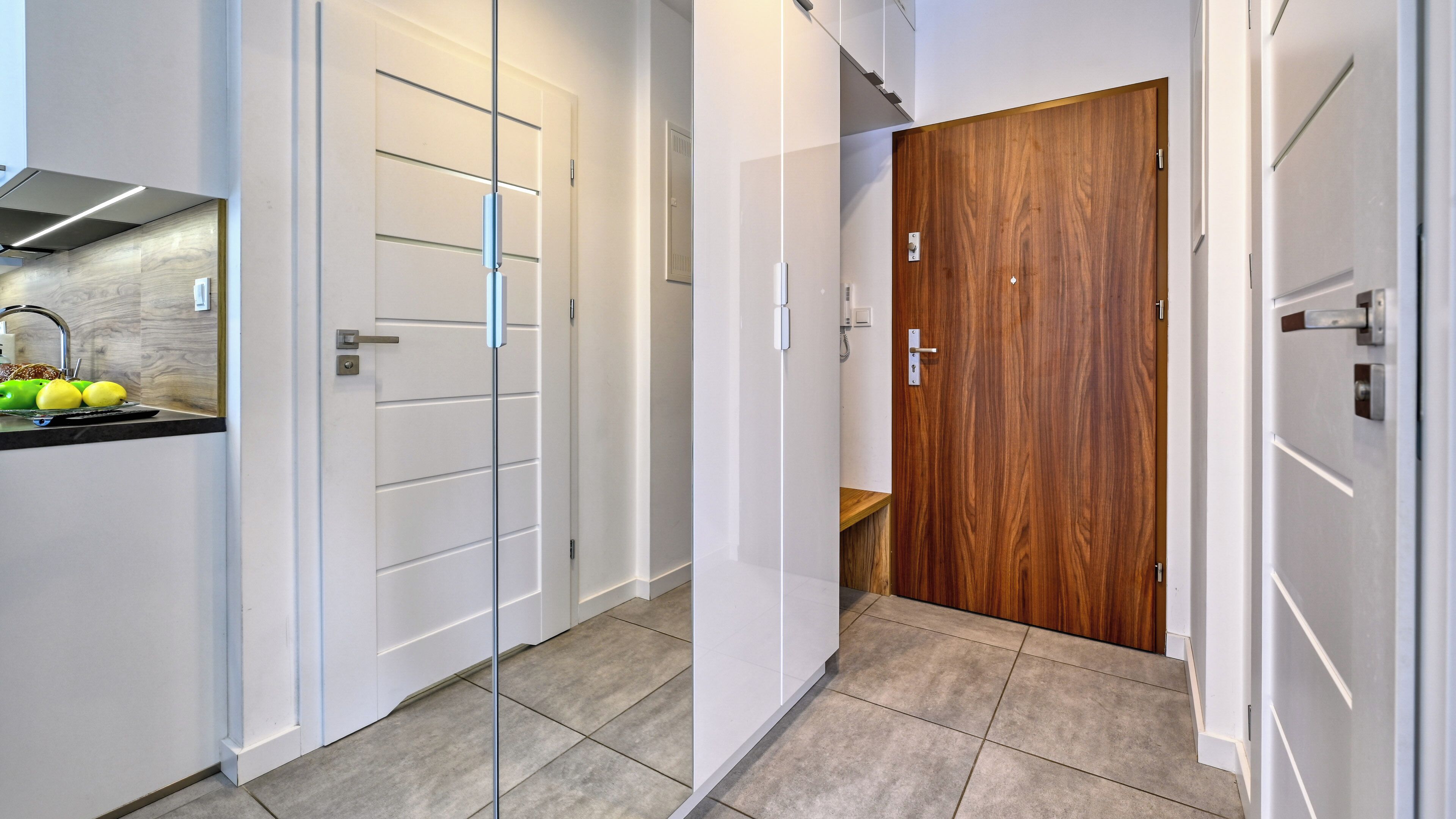 Um corredor estreito com portas brancas, uma porta de madeira e uma área de cozinha com pia e frutas.