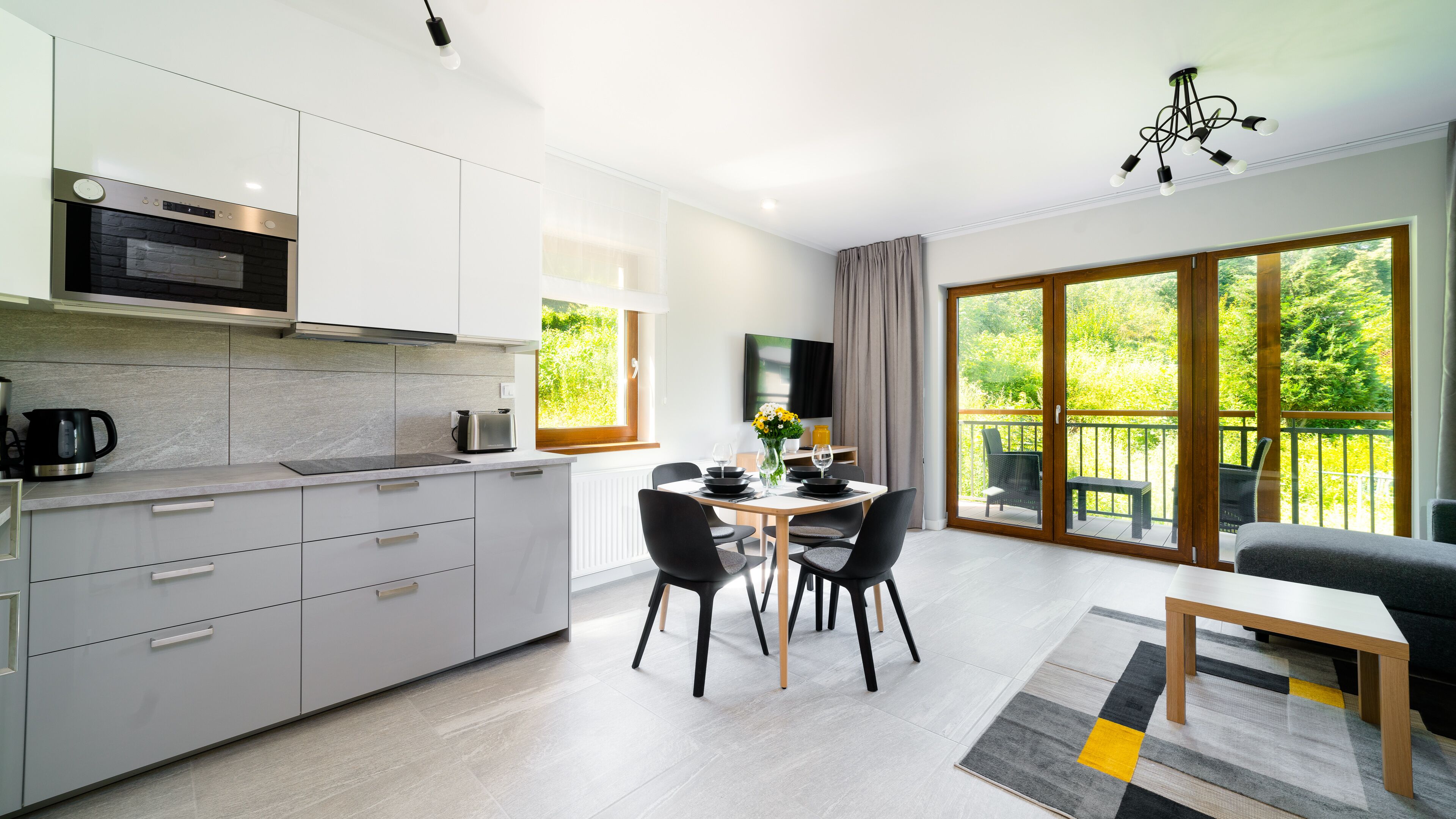 A modern kitchen with a built-in oven, a dining area with a table set for four, and a balcony with a view of greenery.