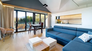 A modern living room with a blue sofa, wooden coffee table, wicker chairs, and a dining table. Large windows offer a view of trees and hills.