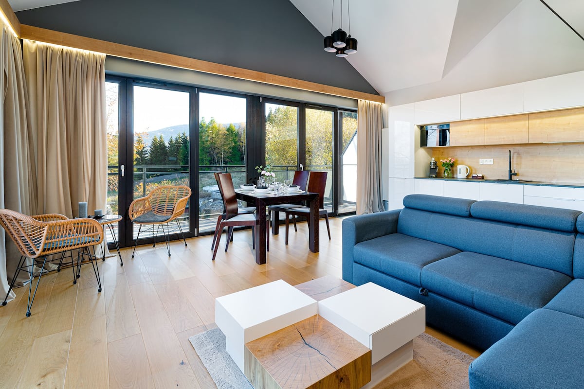 Un salon moderne avec un canapé bleu, une table basse en bois, des fauteuils en osier et une table à manger. De grandes fenêtres offrent une vue sur les arbres et les collines.