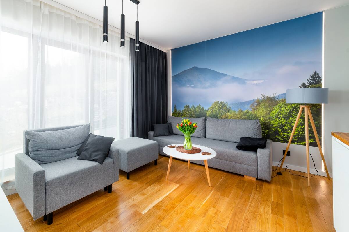 Un salon moderne avec un canapé gris, une table basse ornée d’un vase de fleurs et une grande fenêtre avec des rideaux légers.