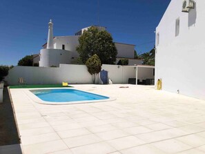 Une piscine avec une terrasse carrelée de blanc, un espace salon couvert et un bâtiment blanc doté d’une cheminée.