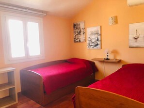 Une petite chambre agréable, dotée de deux lits simples, d’une fenêtre avec des stores et d’œuvres d’art accrochées aux murs.