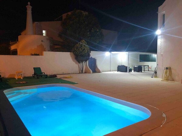 A swimming pool with a tiled deck, a white building with a chimney, and outdoor furniture.