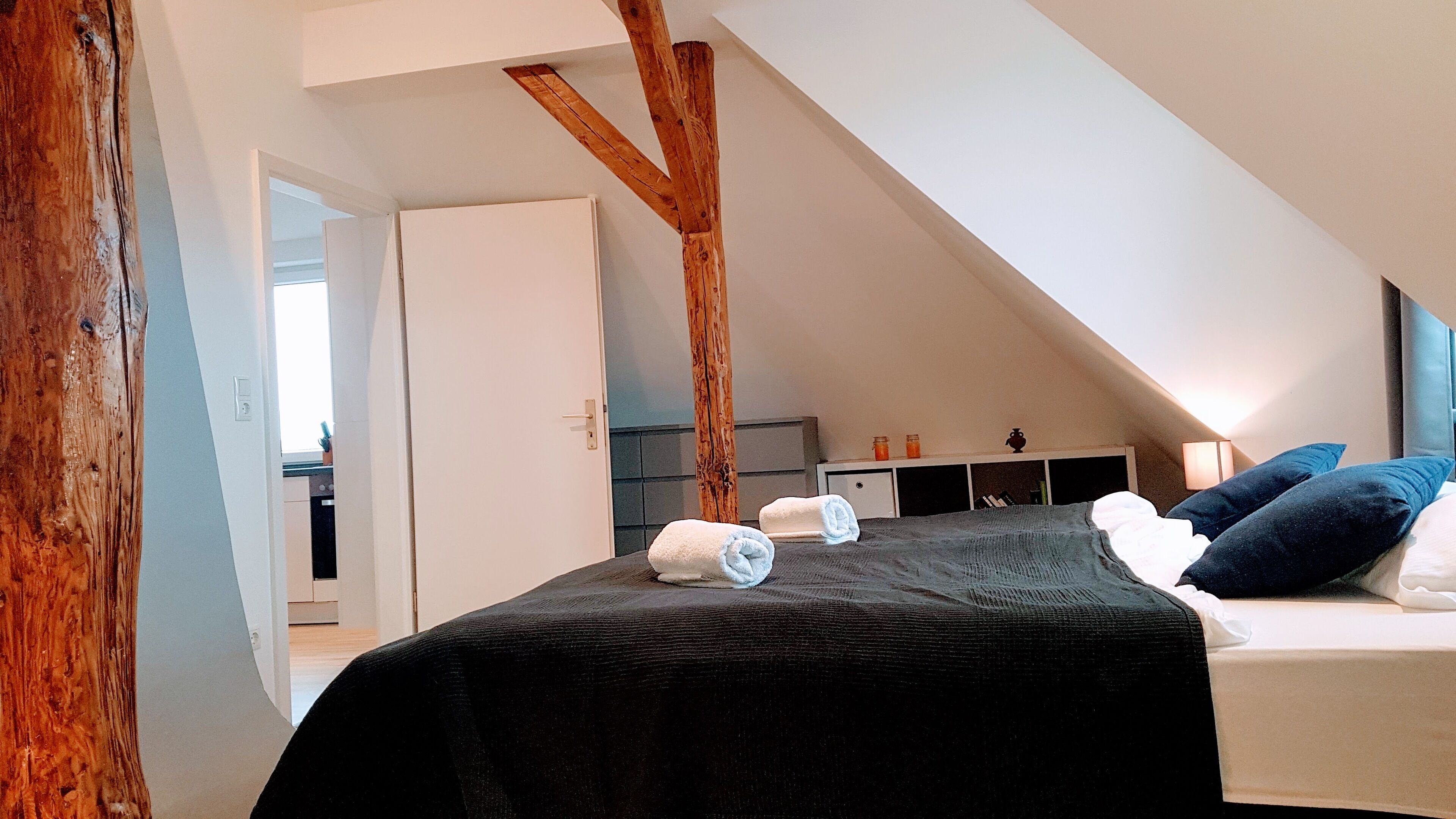 A bedroom with a sloped ceiling, a bed with a dark cover, two rolled towels, a bedside table with a lamp, and a small shelf with items.