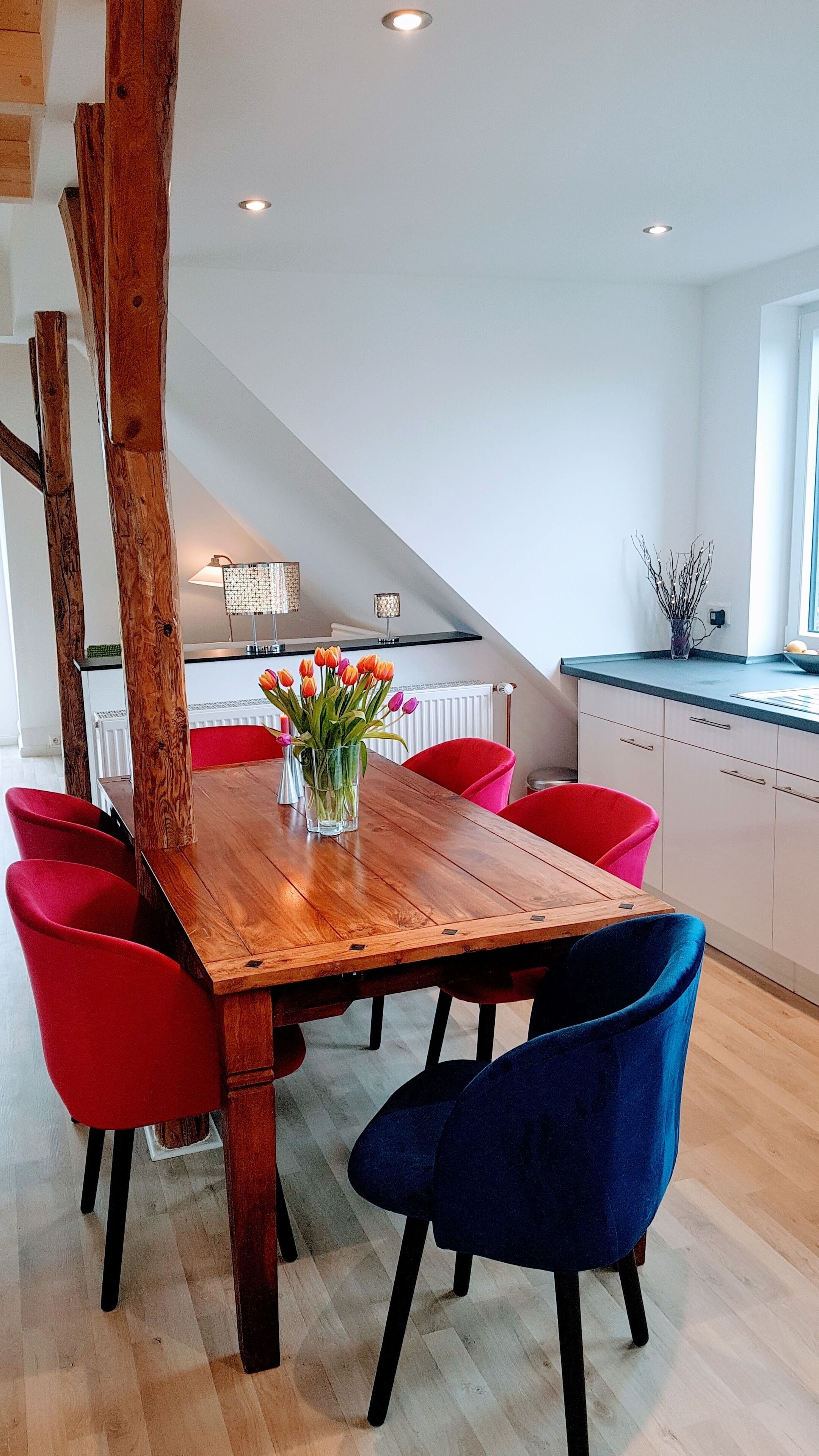 Een eetruimte met een houten tafel, rode en blauwe stoelen en een vaas tulpen.