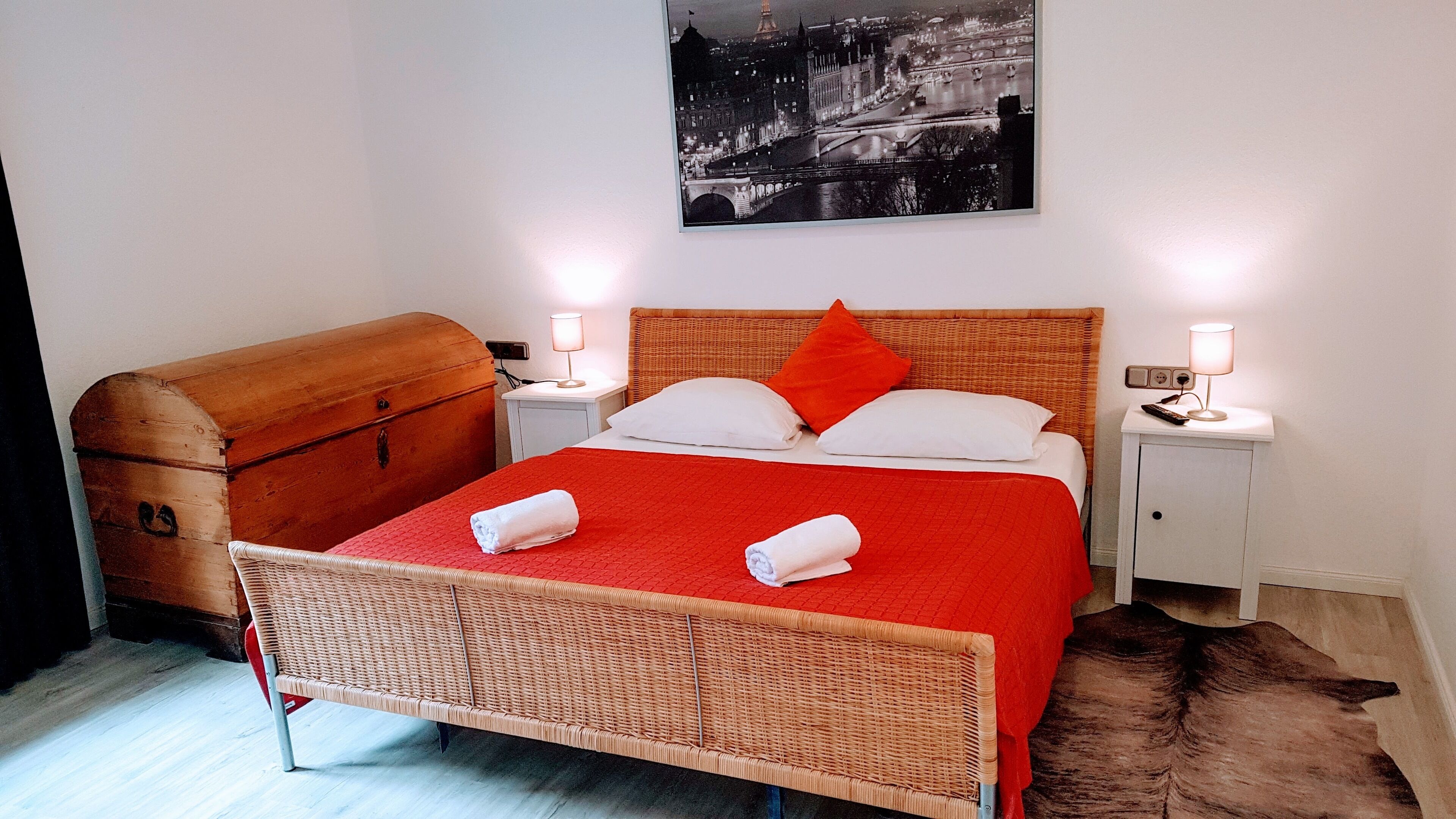 Une chambre d’hôtel avec un lit, une commode en bois, deux tables de chevet avec des lampes et un tableau représentant un paysage urbain accroché au mur.