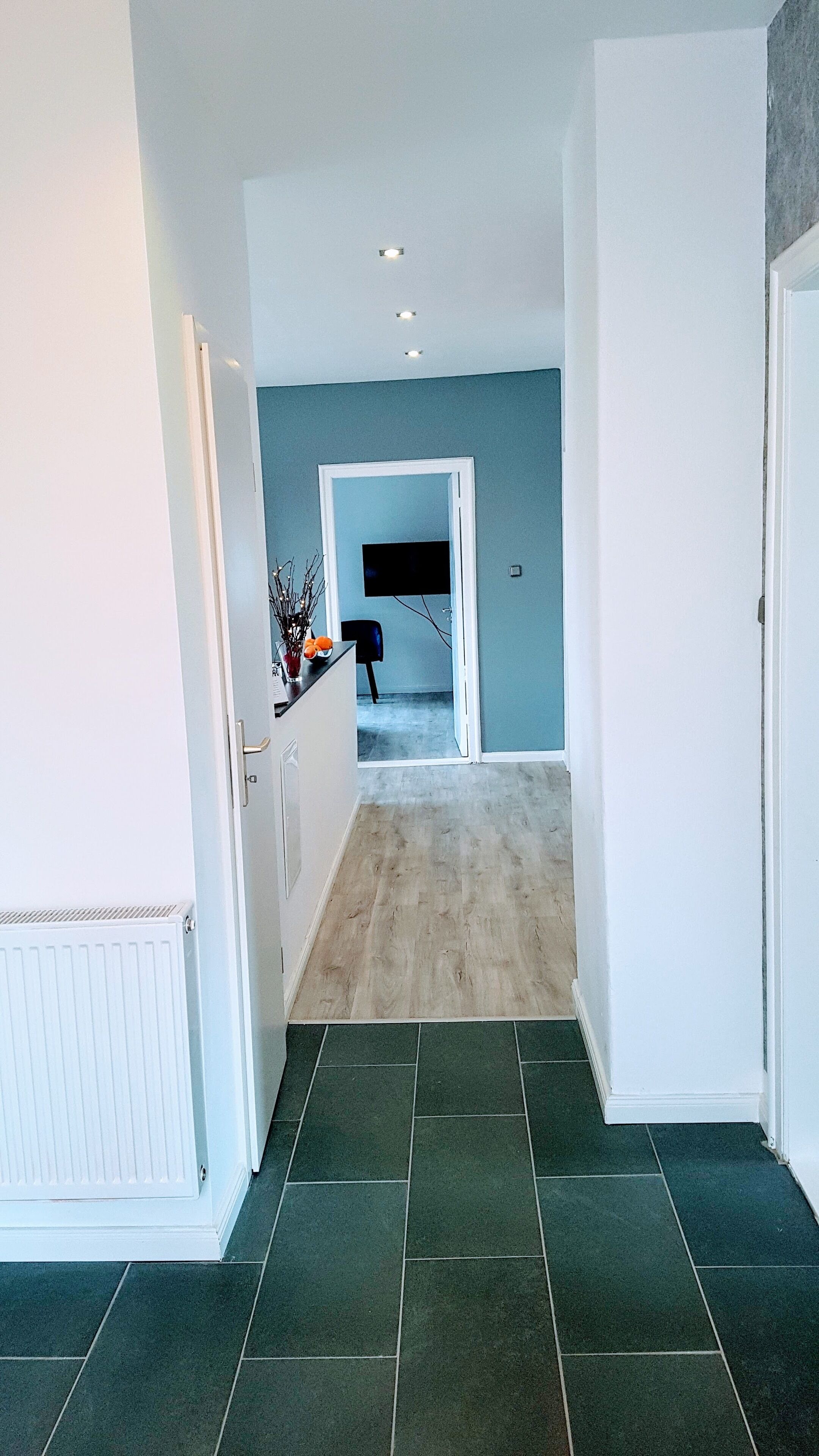 Un couloir avec un mur bleu clair, un plancher en bois et une zone recouverte de carreaux vert foncé.