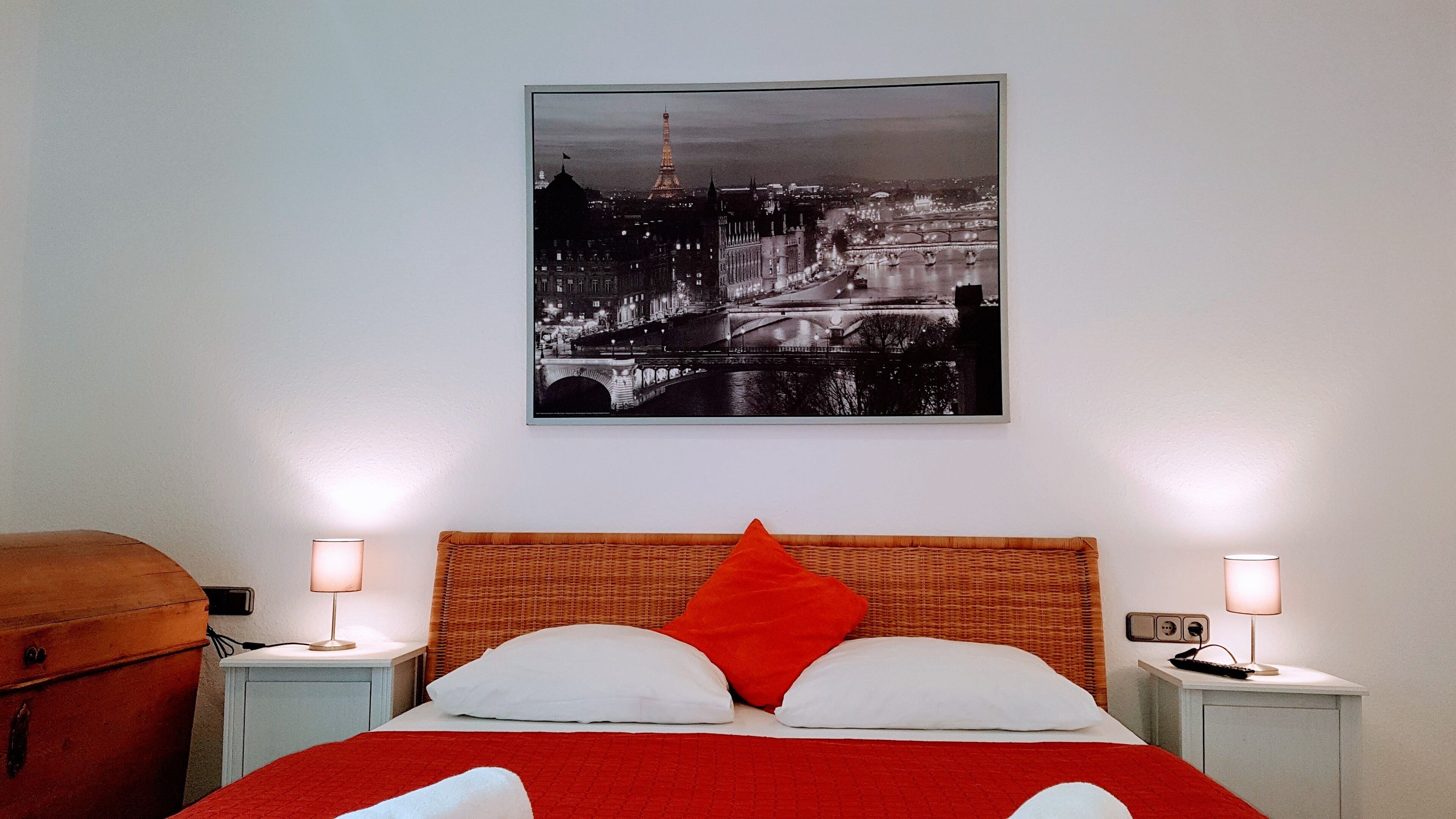 A hotel room with a bed, two bedside tables, a lamp on each table, a red blanket, and a cityscape picture on the wall.