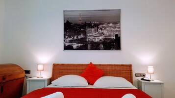 A hotel room with a bed, two bedside tables, a lamp on each table, a red blanket, and a cityscape picture on the wall.