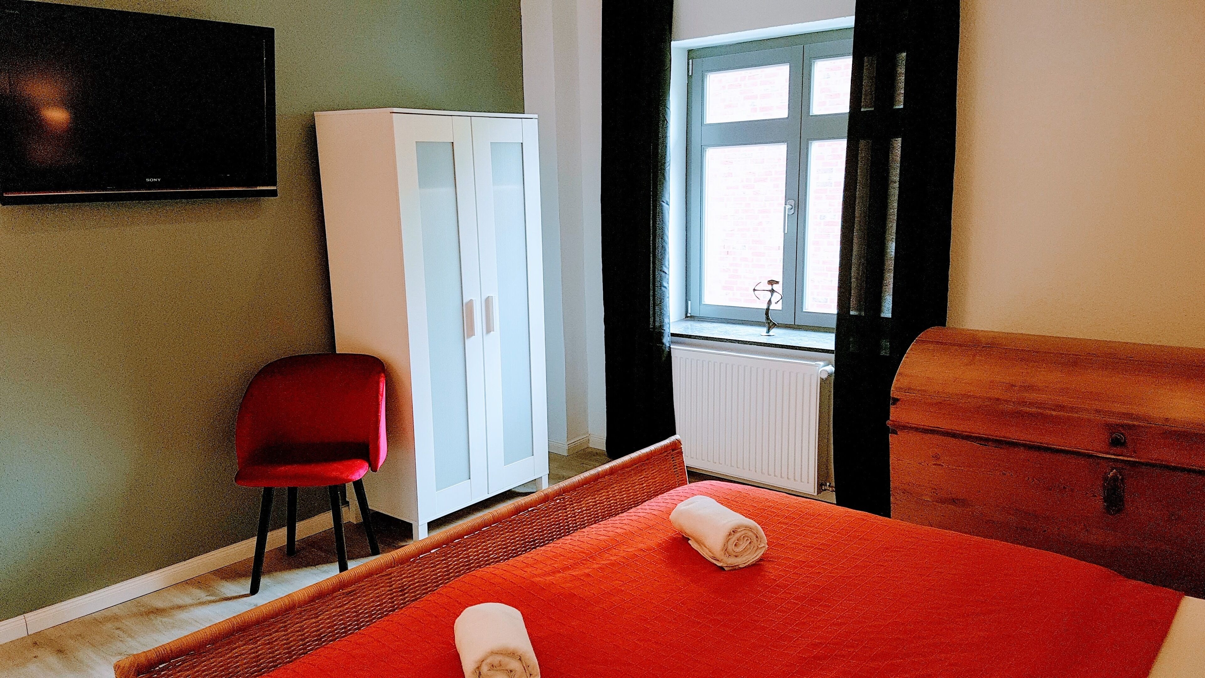 A hotel room with a bed, a red chair, a white wardrobe, and a television on the wall.
