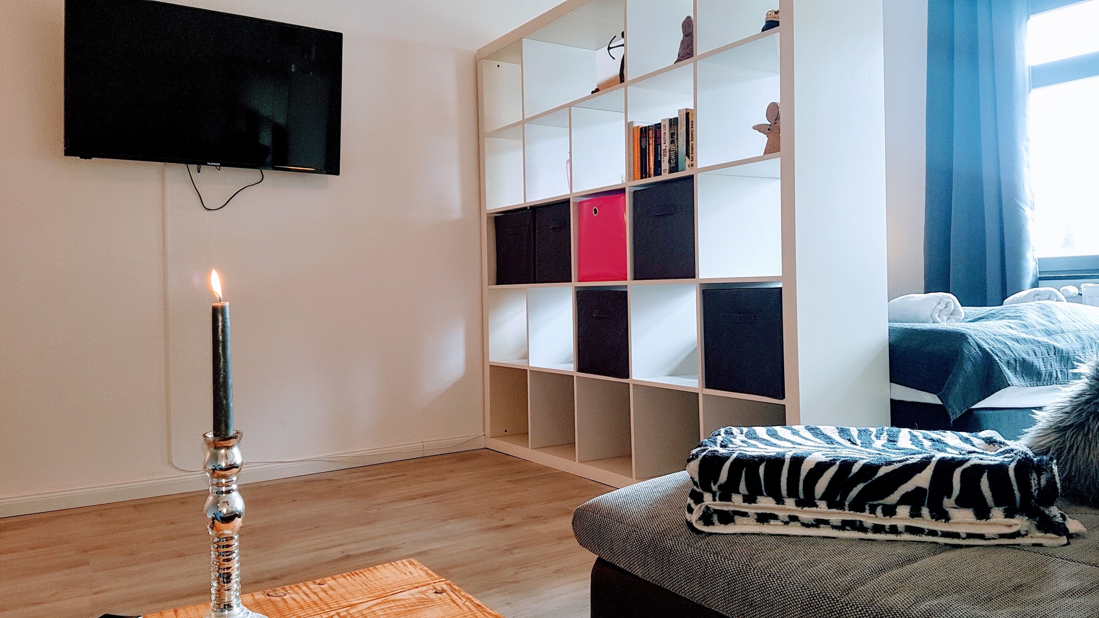 A room with a flat-screen TV mounted on the wall, a bookshelf with various items, a bed with a zebra-patterned blanket, and a lit candle on a wooden table.