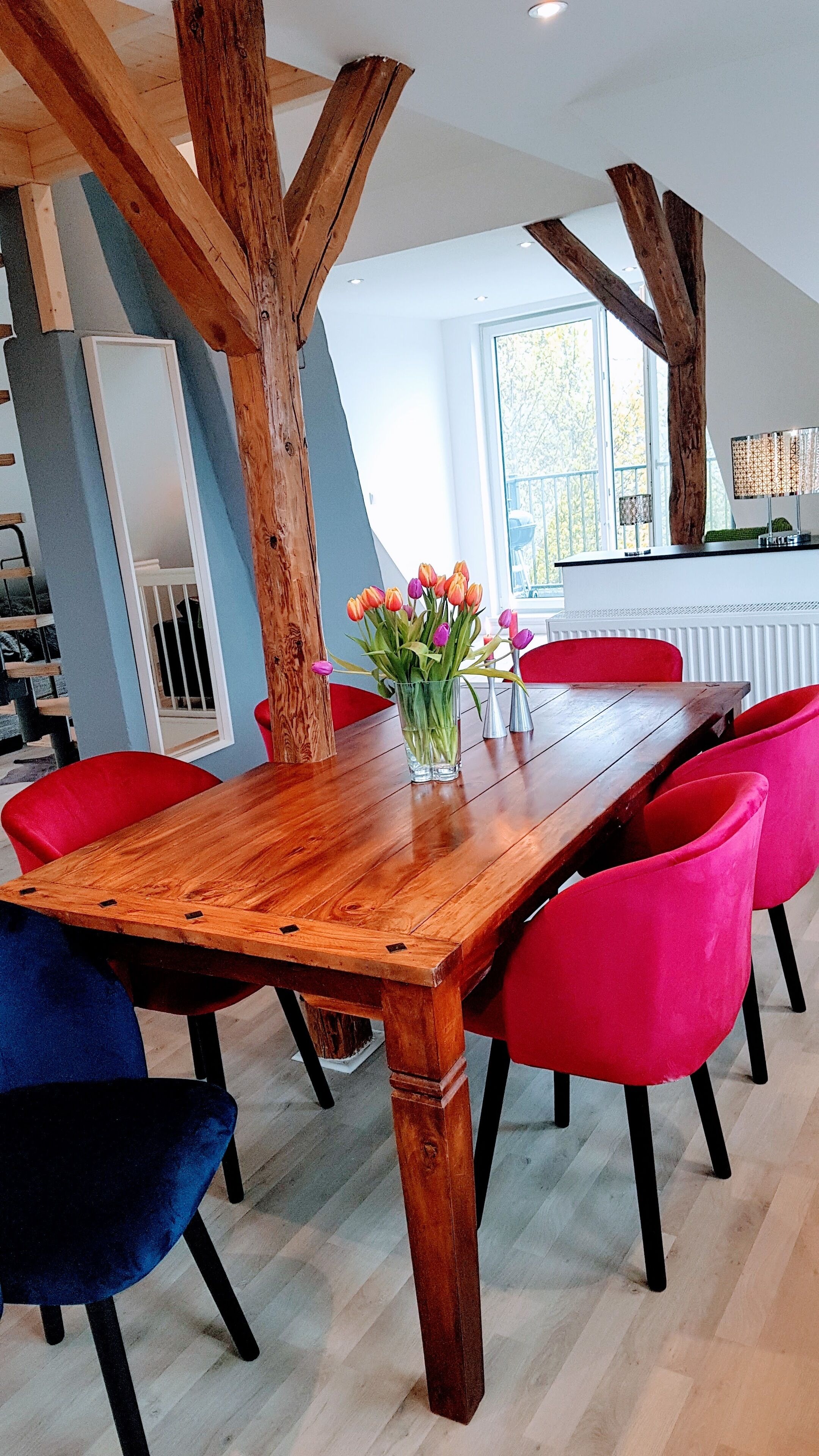 Un comedor con una mesa de madera, sillas rojas y azules, y un florero con flores.