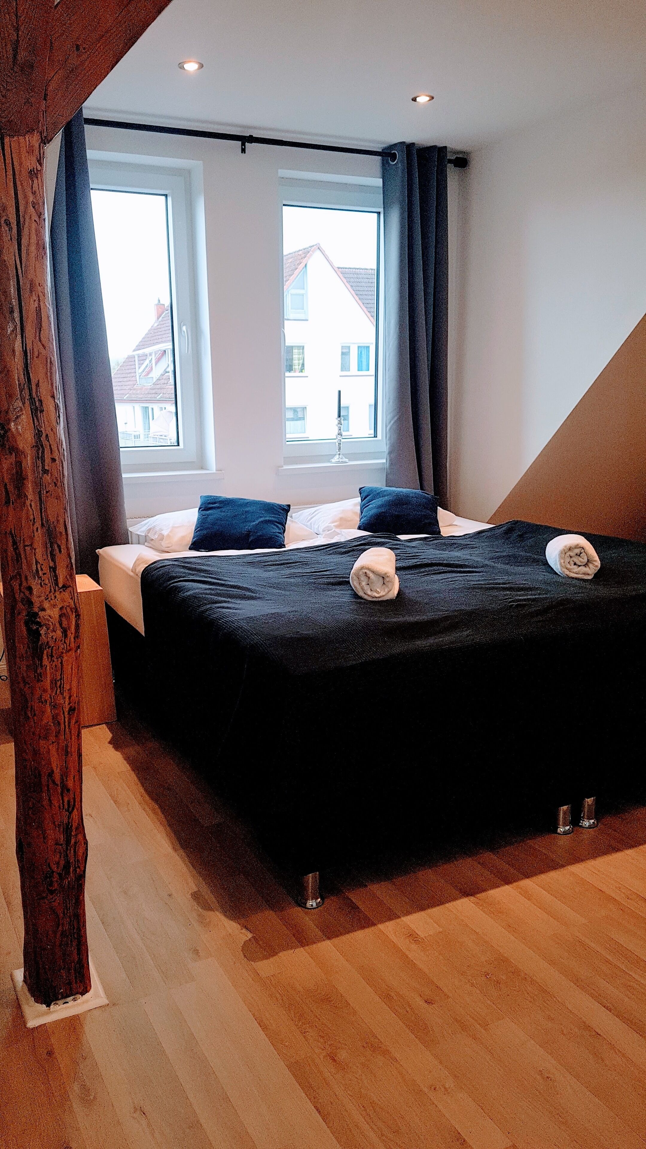 A bedroom with a bed, two windows, and a wooden beam structure.