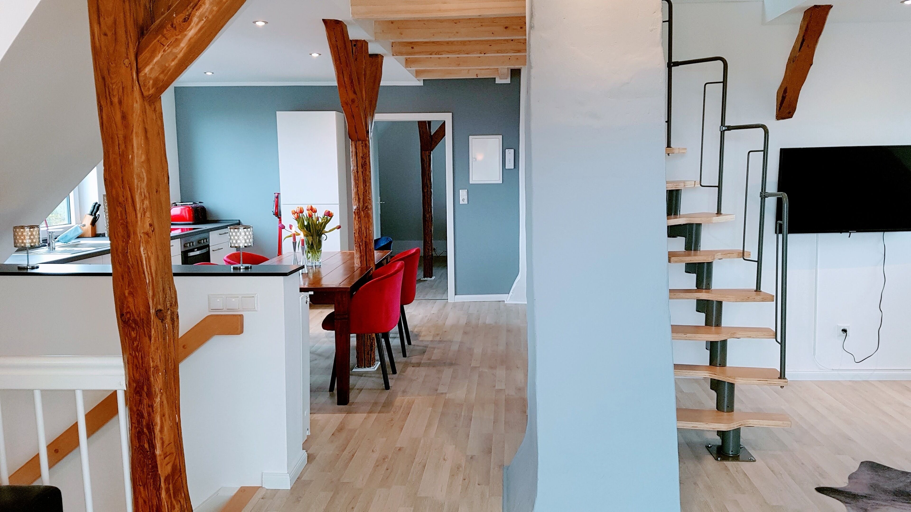Eine moderne Küche mit Holzbalken, ein Essbereich mit roten Stühlen und eine Treppe mit Fernseher.